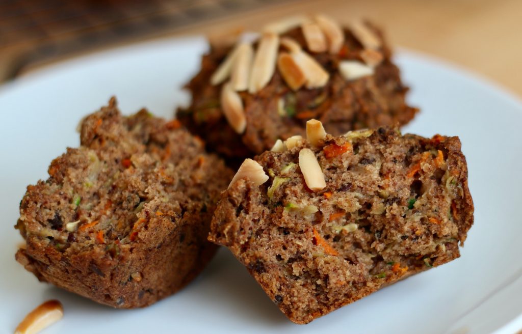 Two zucchini carrot muffins on a small white plate. One of the muffins is split in half so you can see the texture of the center.