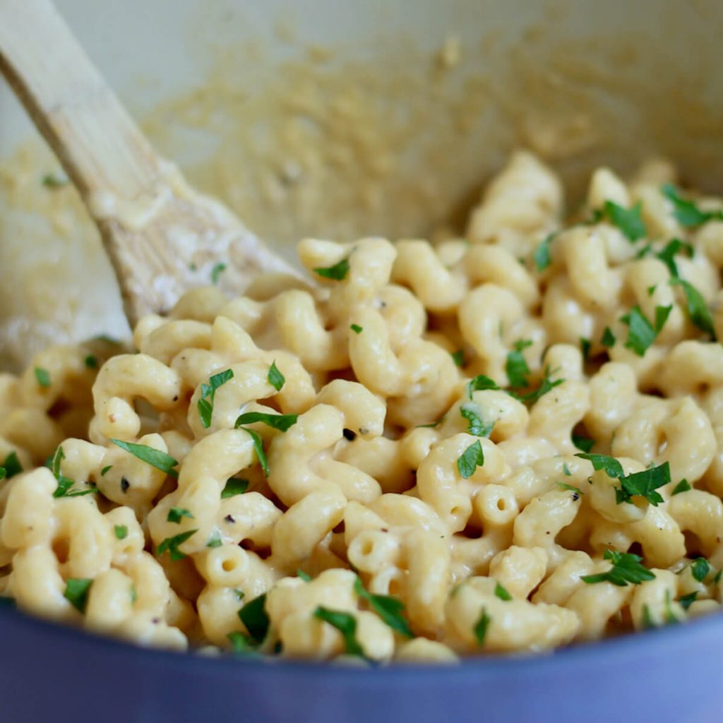 A lavender dutch oven filled with creamy stovetop macaroni and cheese. The pasta is garnished with fresh parsley and there is a wooden spoon sticking out of the pot in the background.