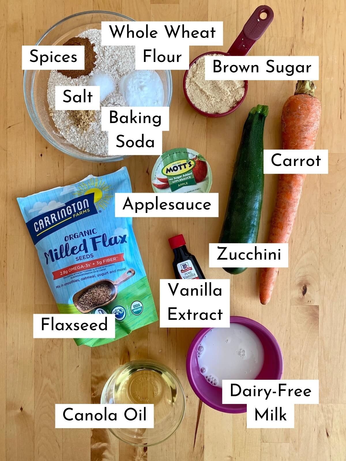 All of the ingredients to make zucchini carrot muffins laid out on a counter. There is text over each ingredient labeling what it is. The ingredients are whole wheat flour, brown sugar, baking soda, salt, spices, applesauce, zucchini, carrots, flaxseed, vanilla extract, canola oil, and dairy-free milk.