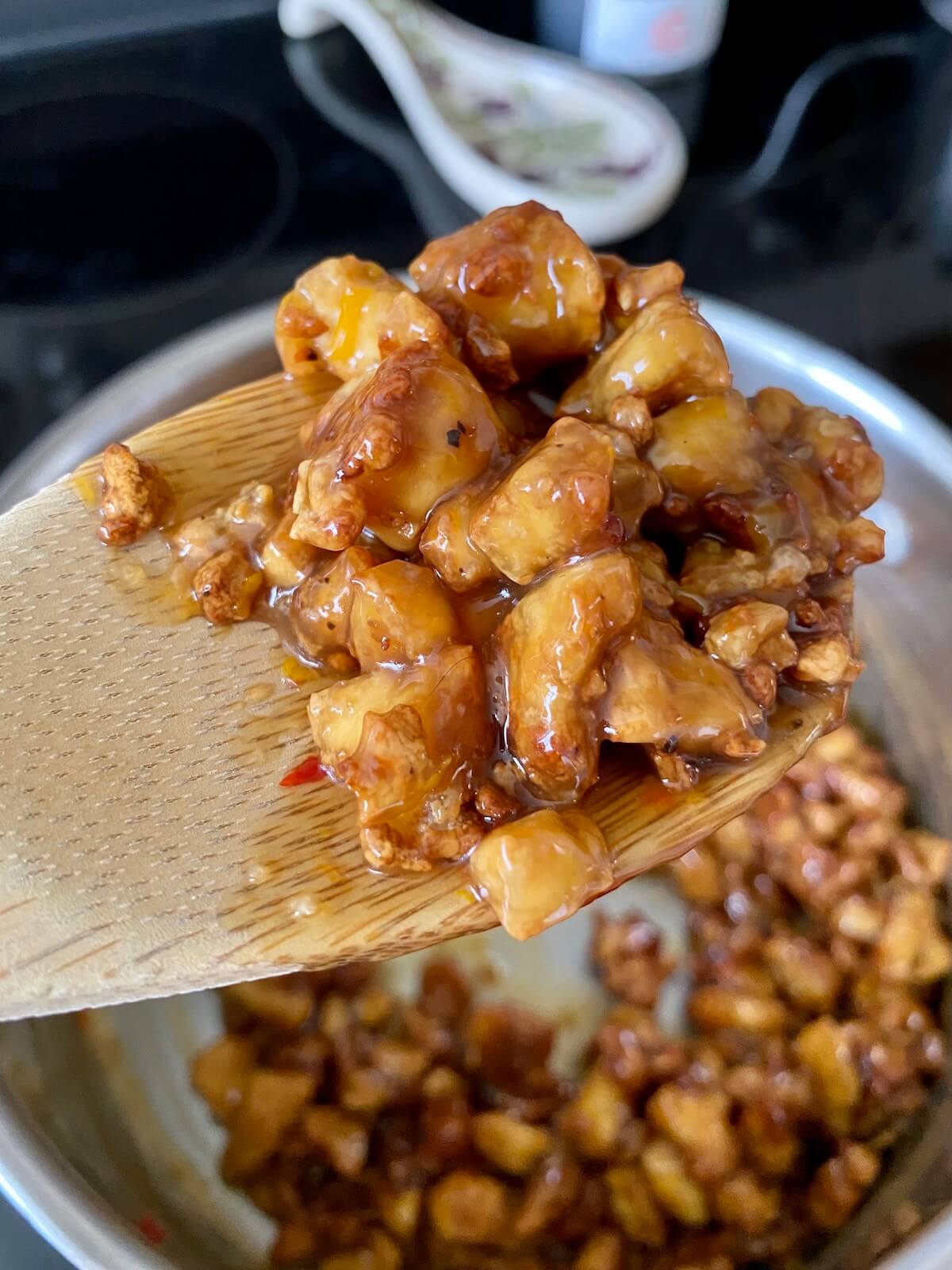A wooden spoon holds up some of the finished baked orange tofu. The saucepan filled with the rest of the tofu is in the background.
