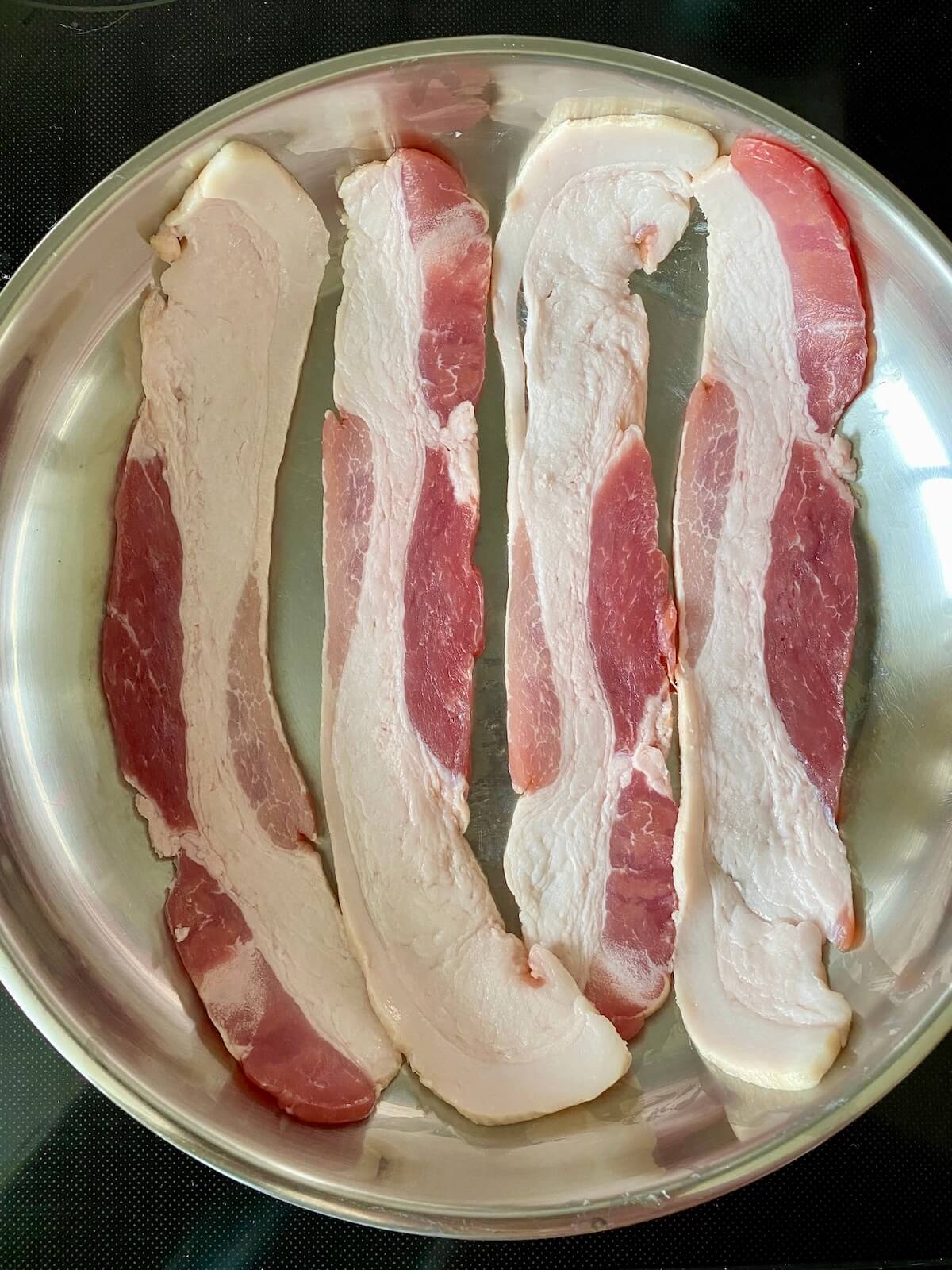 Four strips of raw bacon in a pan on the stove.