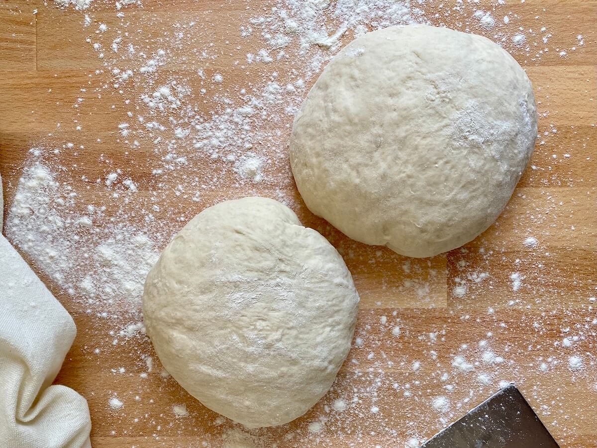 Two balls of 72-hour pizza dough sitting on a lightly floured countertop next to a white kitchen towel and a bench scraper.