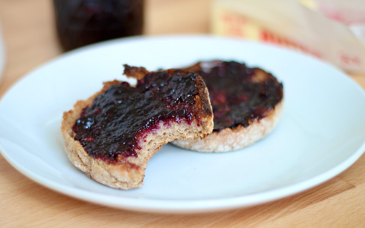 A whole wheat English muffin with a bite taken out of it sitting on a white plate. On the English muffin is some butter and blueberry jam.