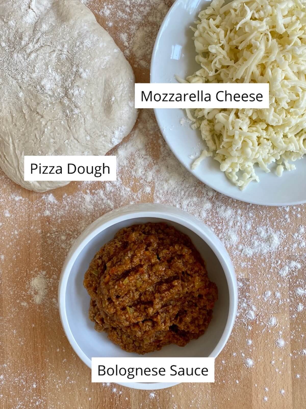 A bowl of bolognese sauce, a plate of shredded mozzarella, and a ball of pizza dough on a lightly floured surface.