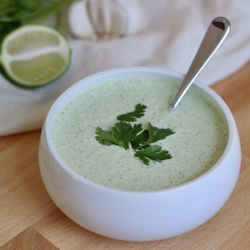 A white bowl filled with cilantro garlic sauce. It is garnished with a few leaves of cilantro and a silver spoon is inside the bowl.