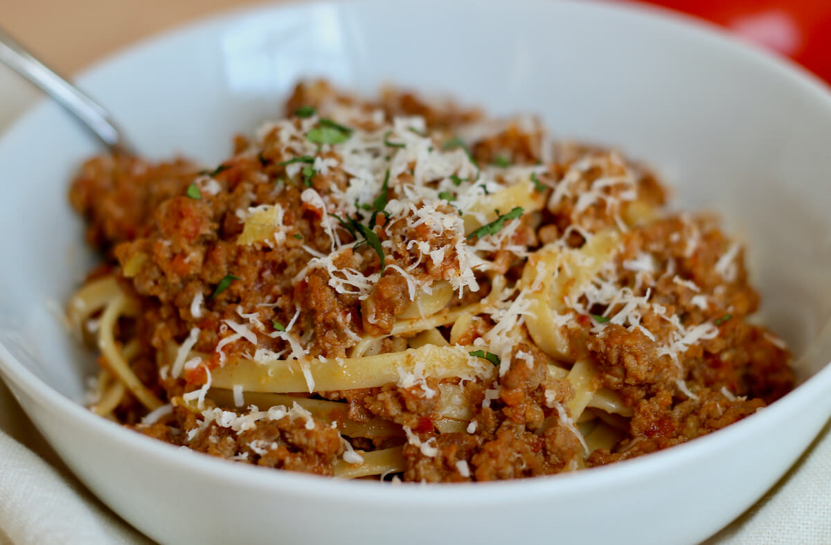 A white bowl filled with pasta dressed in creamy bolognese sauce and garnished with parmesan cheese and parsley. In the background, a fork is sticking out of the bowl.