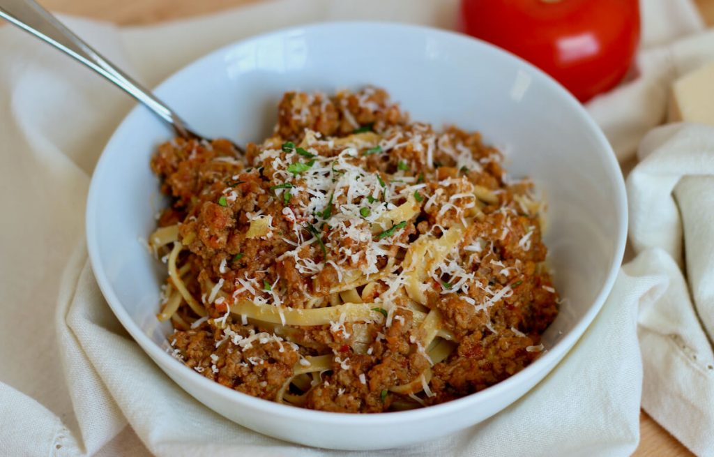 A white bowl filled with fettuccini dressed in creamy bolognese sauce. The dish is garnished with grated parmesan cheese and parsley. In the background, a fork is sticking out of the bowl.