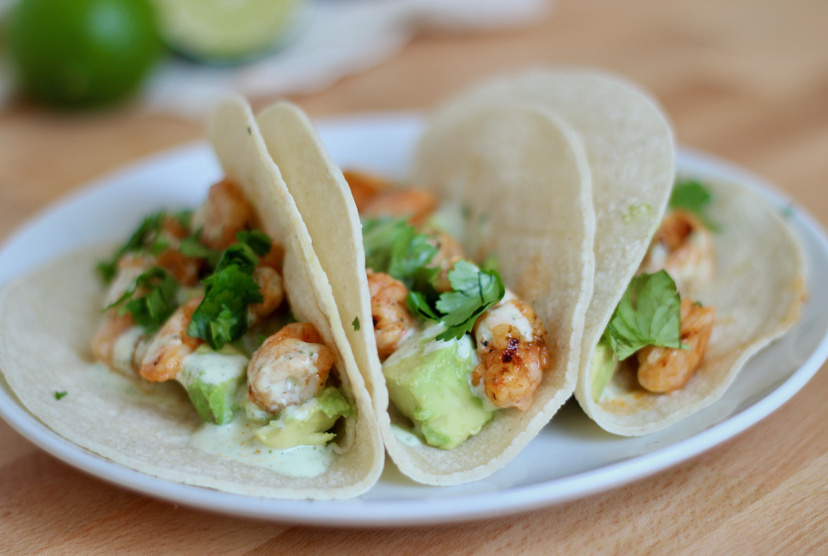 Three honey chipotle shrimp tacos on a white plate. The tacos are garnished with cilantro garlic sauce and diced avocado.