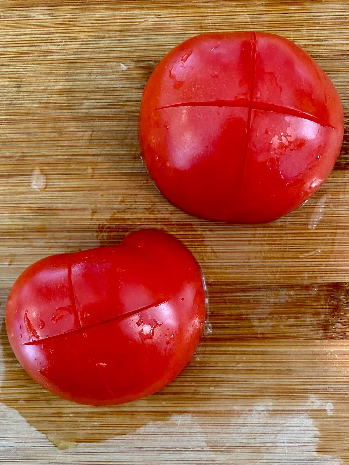 A tomato cut-side-down on a bamboo cutting board. The back of each tomato is scored with an X.