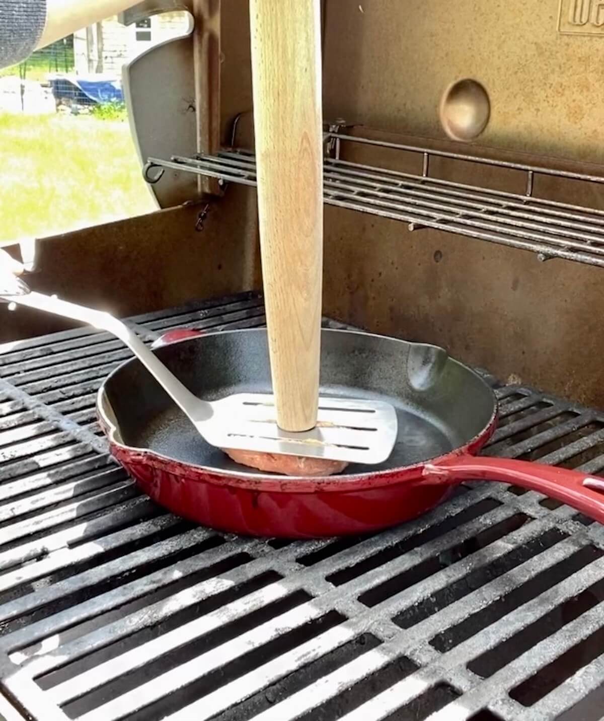 A turkey burger being smashed into a cast iron skillet on the grill using a spatula and wooden rolling pin.