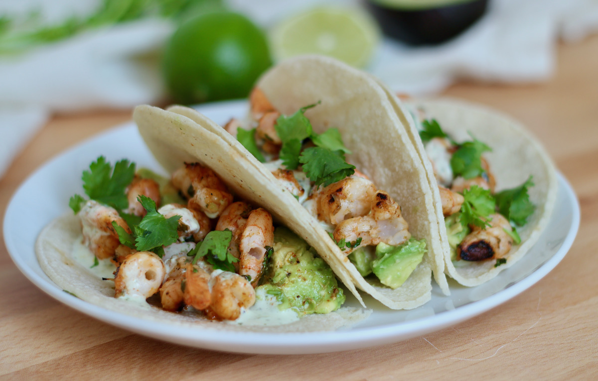 Three tequila lime shrimp tacos garnished with cilantro, avocado, and cilantro garlic sauce are served on a white plate.