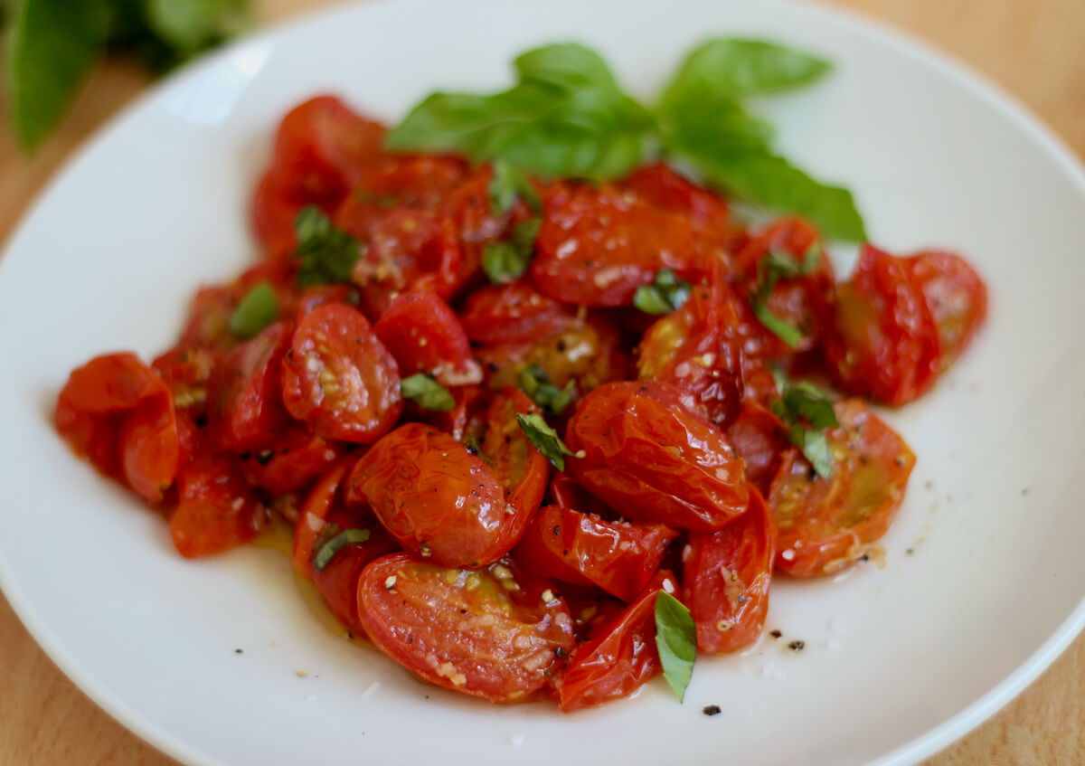 Roasted air fryer tomatoes on a white plate. They are drizzled with extra virgin olive oil and sprinkled with kosher salt, freshly cracked black pepper, and fresh basil.