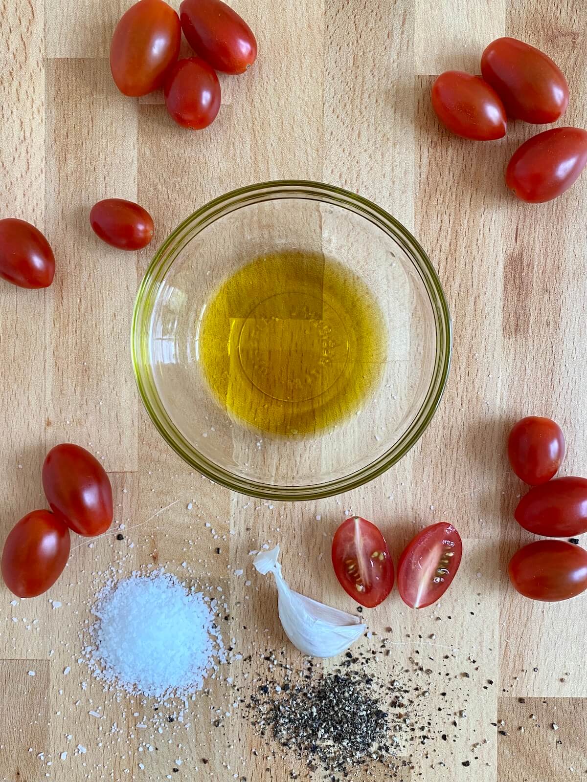 The ingredients to make air fryer cherry tomatoes are laid out on a butcher block countertop. They include cherry tomatoes, extra virgin olive oil, garlic, salt, and pepper.