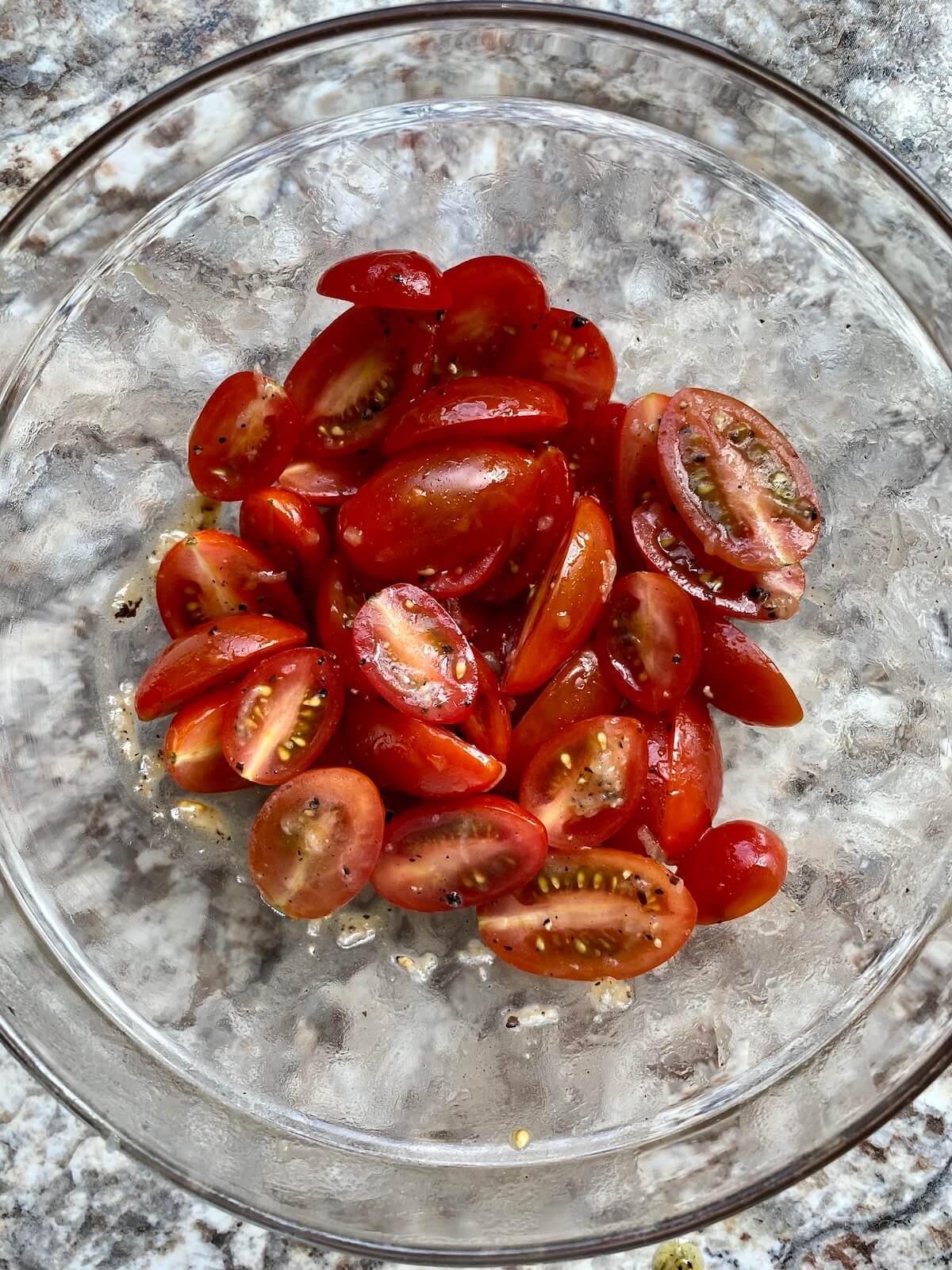 A clear glass bowl filled with halved cherry tomatoes coated with olive oil and seasonings.
