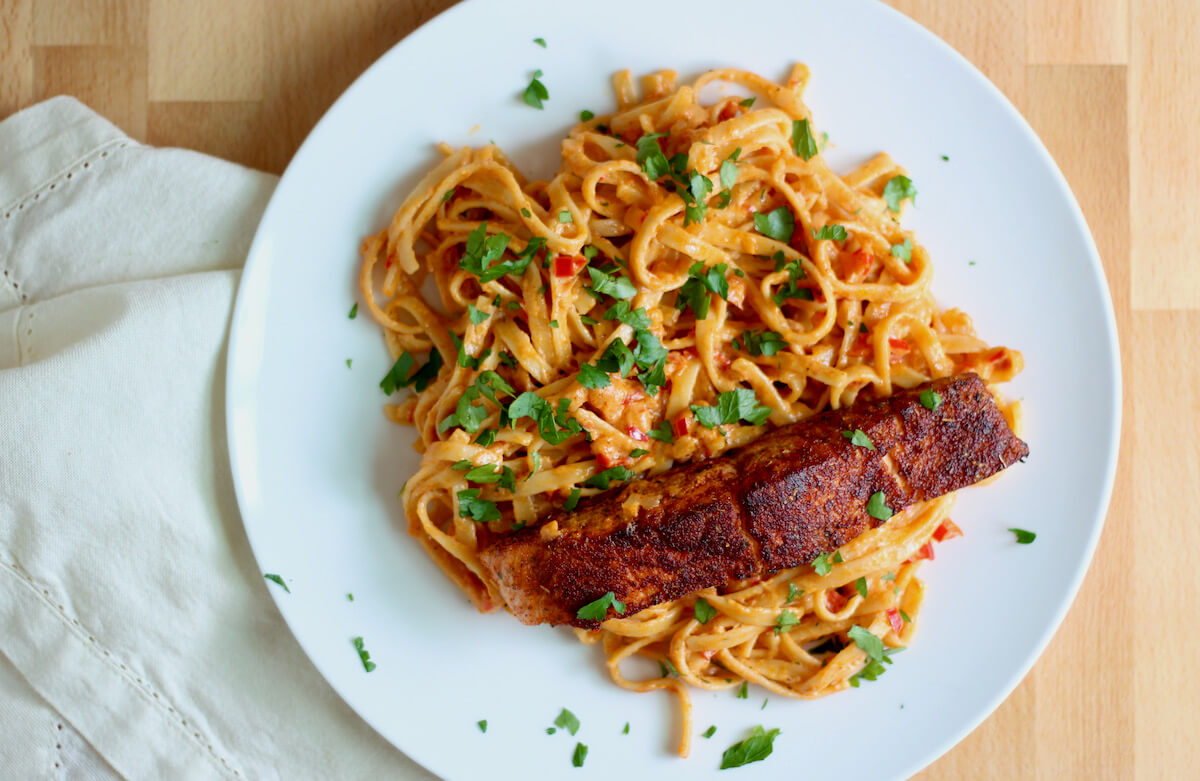 A large white plate filled with creamy Cajun alfredo pasta. The pasta is topped with a fillet of blackened Cajun salmon and garnished with chopped fresh parsley. Next to the plate is a white cloth napkin.