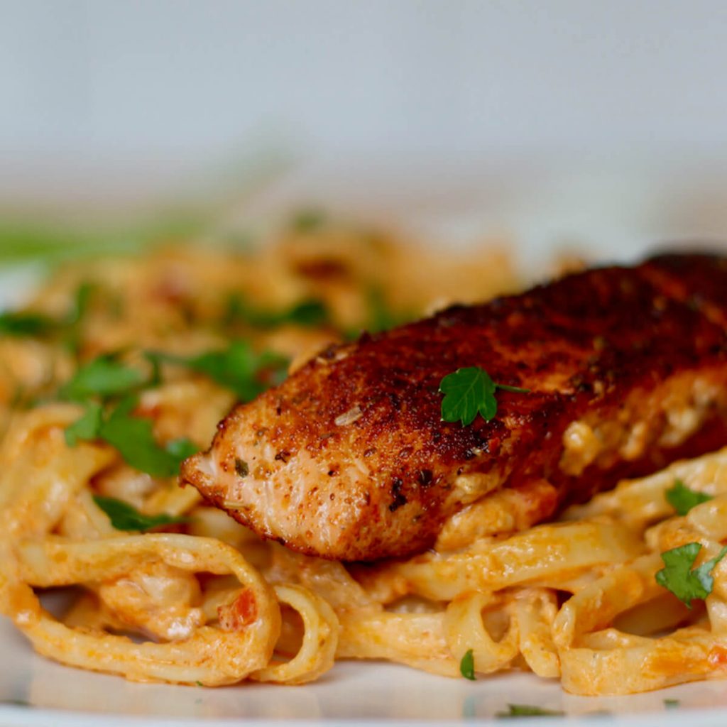 A piece of blackened Cajun salmon on top of a bed of Cajun-spiced fettuccini Alfredo pasta. The white plate is garnished with fresh chopped parsley.