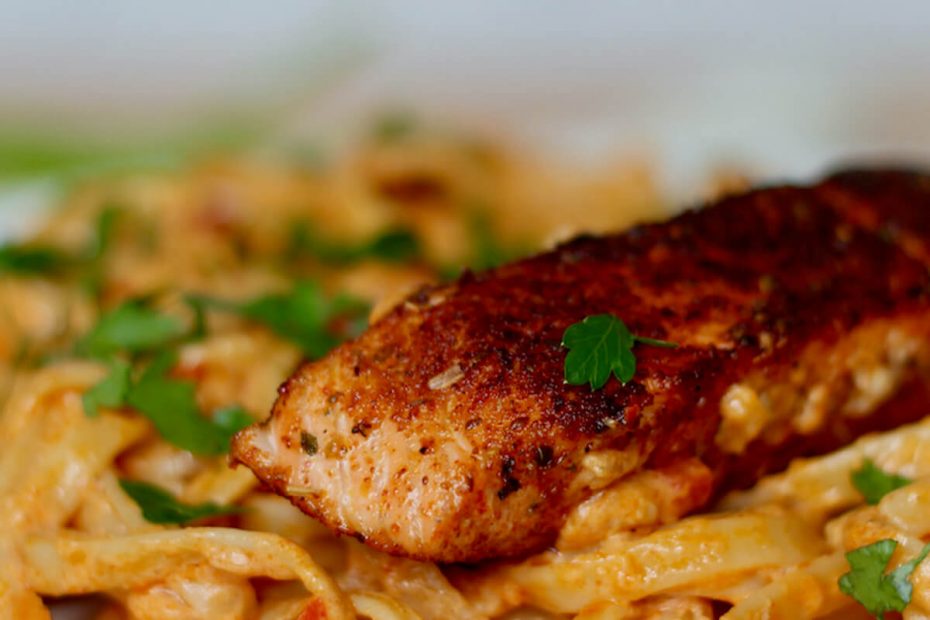 A piece of blackened Cajun salmon on top of a bed of Cajun-spiced fettuccini Alfredo pasta. The white plate is garnished with fresh chopped parsley.
