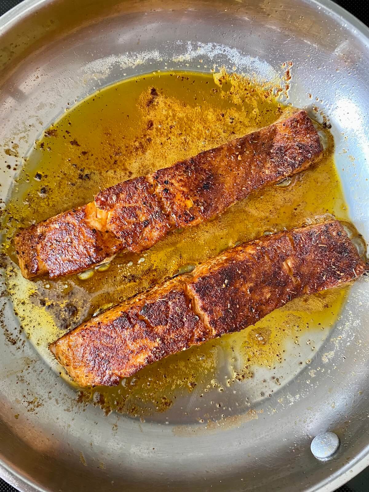 Two fillets of blackened salmon cooking in a stainless steel pan.