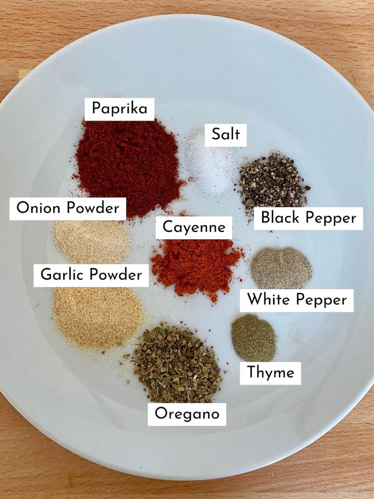 All of the ingredients to make homemade Cajun seasoning displayed on a small, white plate. Each ingredient is labeled with black text on a white background.