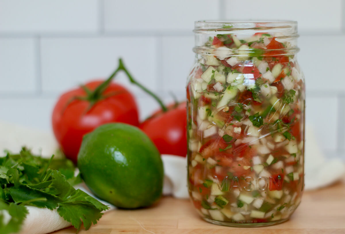 A Ball mason jar filled with cucumber pico de gallo. Out of focus in the background is two tomatoes on the vine, a lime, and some cilantro.