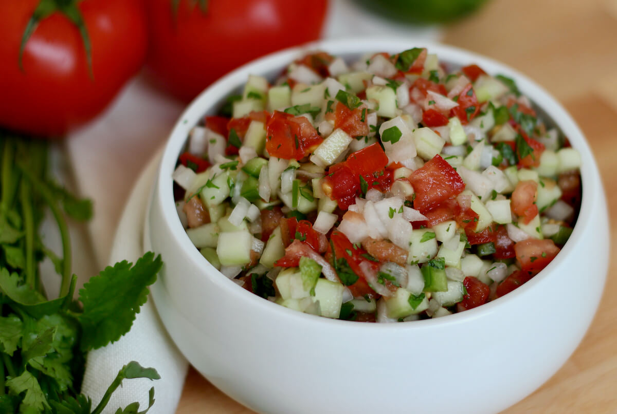 A white bowl brimming with cucumber salsa fresca. Out of focus in the background is two tomatoes and some fresh cilantro.