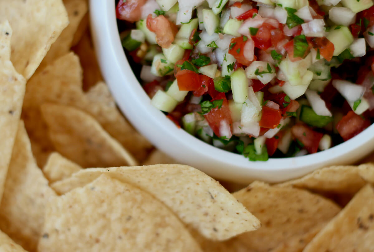 A white bowl filled with cucumber pico de gallo surrounded by corn tortilla chips.