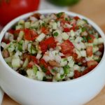 A small white serving bowl filled with cucumber pico de gallo. Out of focus in the background, you can see part of a tomato and a lime.