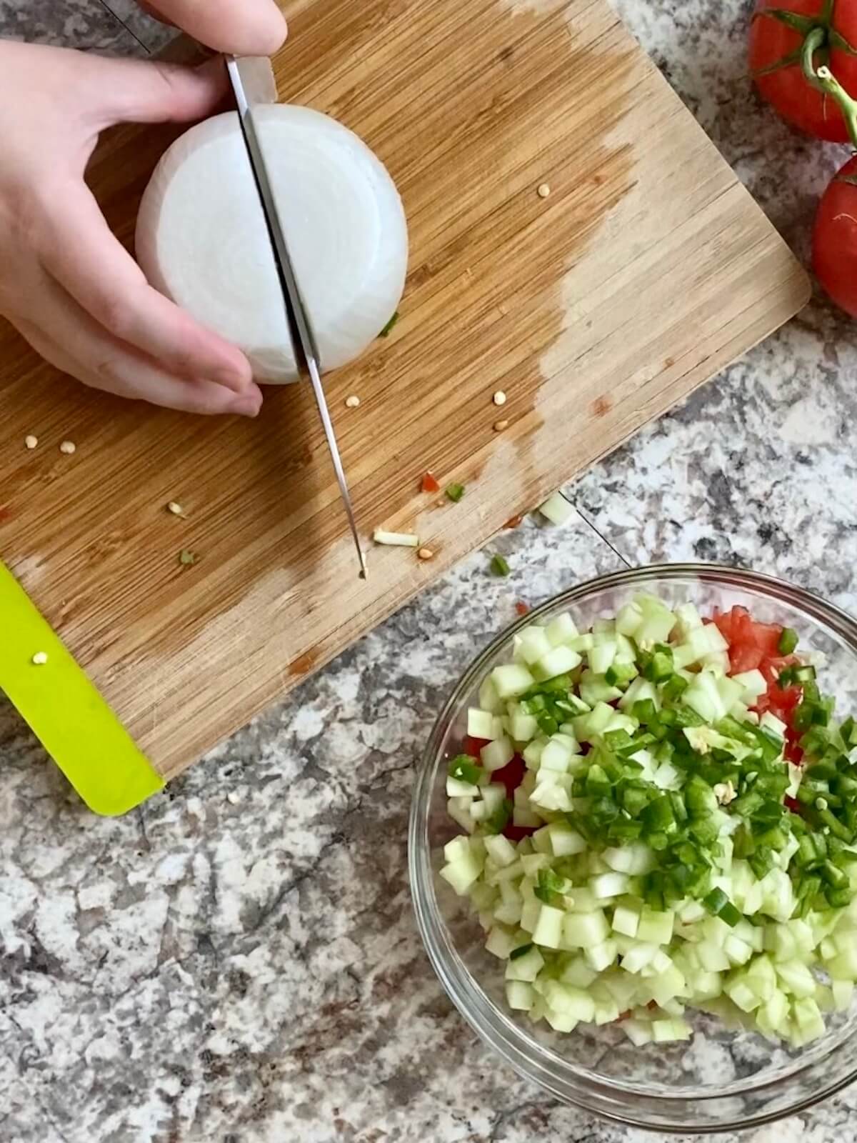 A knife slicing through a white onion on a bamboo cutting board. In front of the cutting board is a glass bowl filled with already chopped vegetables.