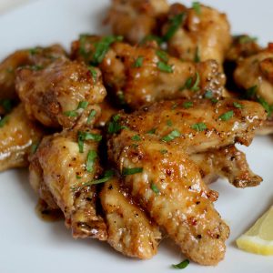 A white plate filled with crispy oven-baked honey lemon pepper wings. The chicken wings are garnished with fresh chopped parsley and served with slices of lemon.