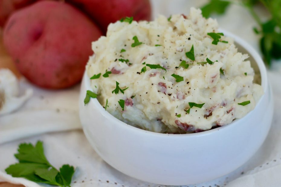 A white bowl overflowing with garlic mashed red skin potatoes. They are garnished with fresh chopped parsley and cracked black pepper.