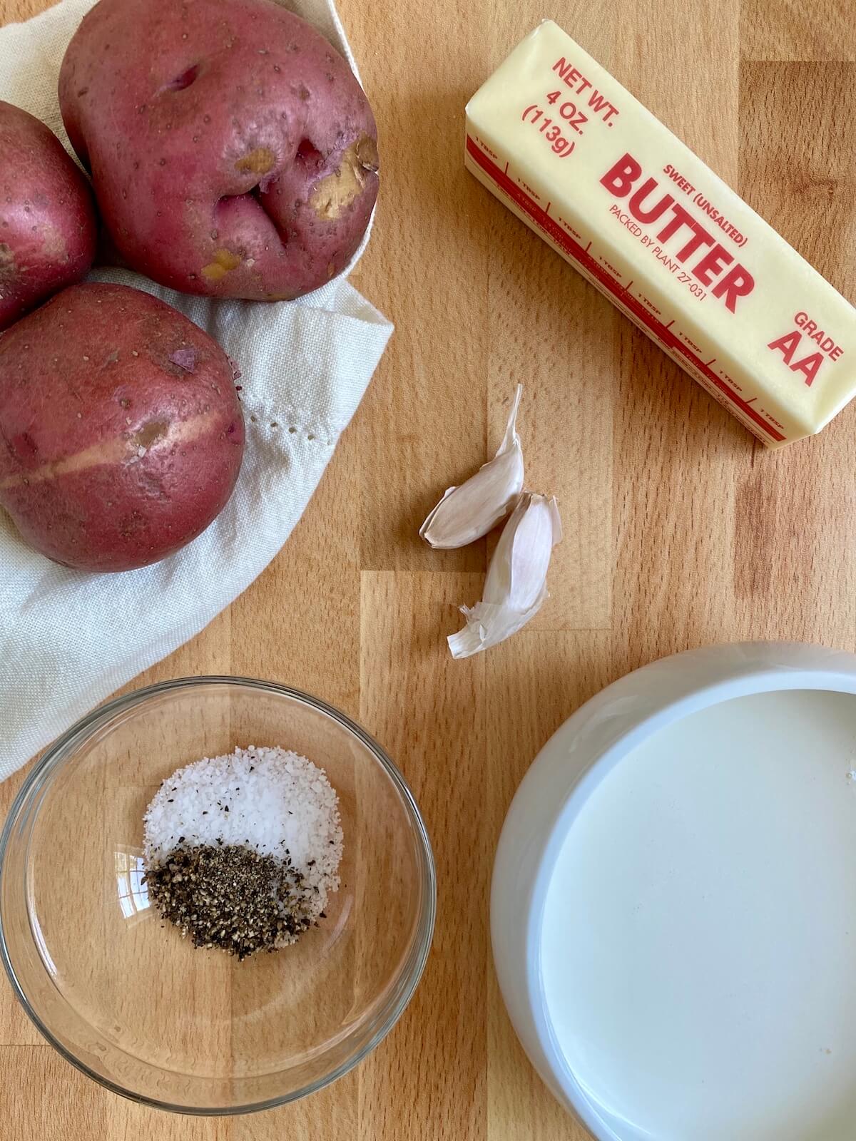 Three whole raw red potatoes, a stick of butter, two unpeeled cloves of garlic, a clear glass bowl with kosher salt and black pepper, and a white bowl filled with light cream are displayed on a butcher block countertop.