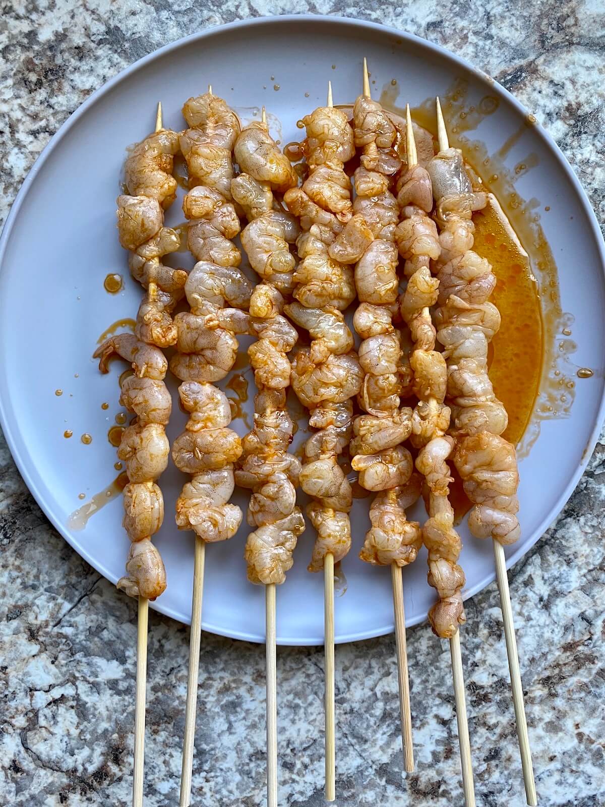 Seven skewers of marinated, raw shrimp on a gray plate.
