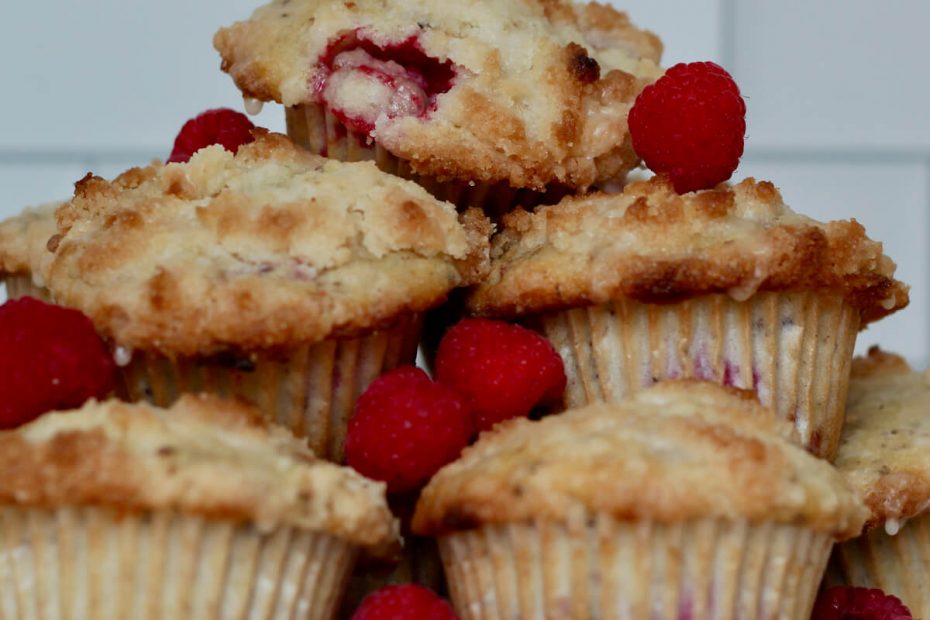 Vegan raspberry muffins stacked on top of one another on a white plate. The muffins are drizzled with a lemon glaze and there are fresh raspberries placed carefully on top of the muffins.
