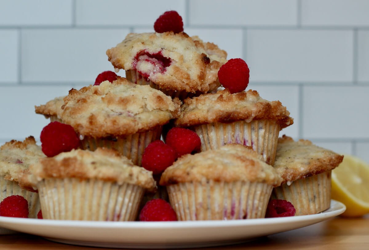 Vegan raspberry muffins stacked on top of each other on a white plate. The stack of muffins is drizzled with a lemon glaze and adorned with fresh raspberries.