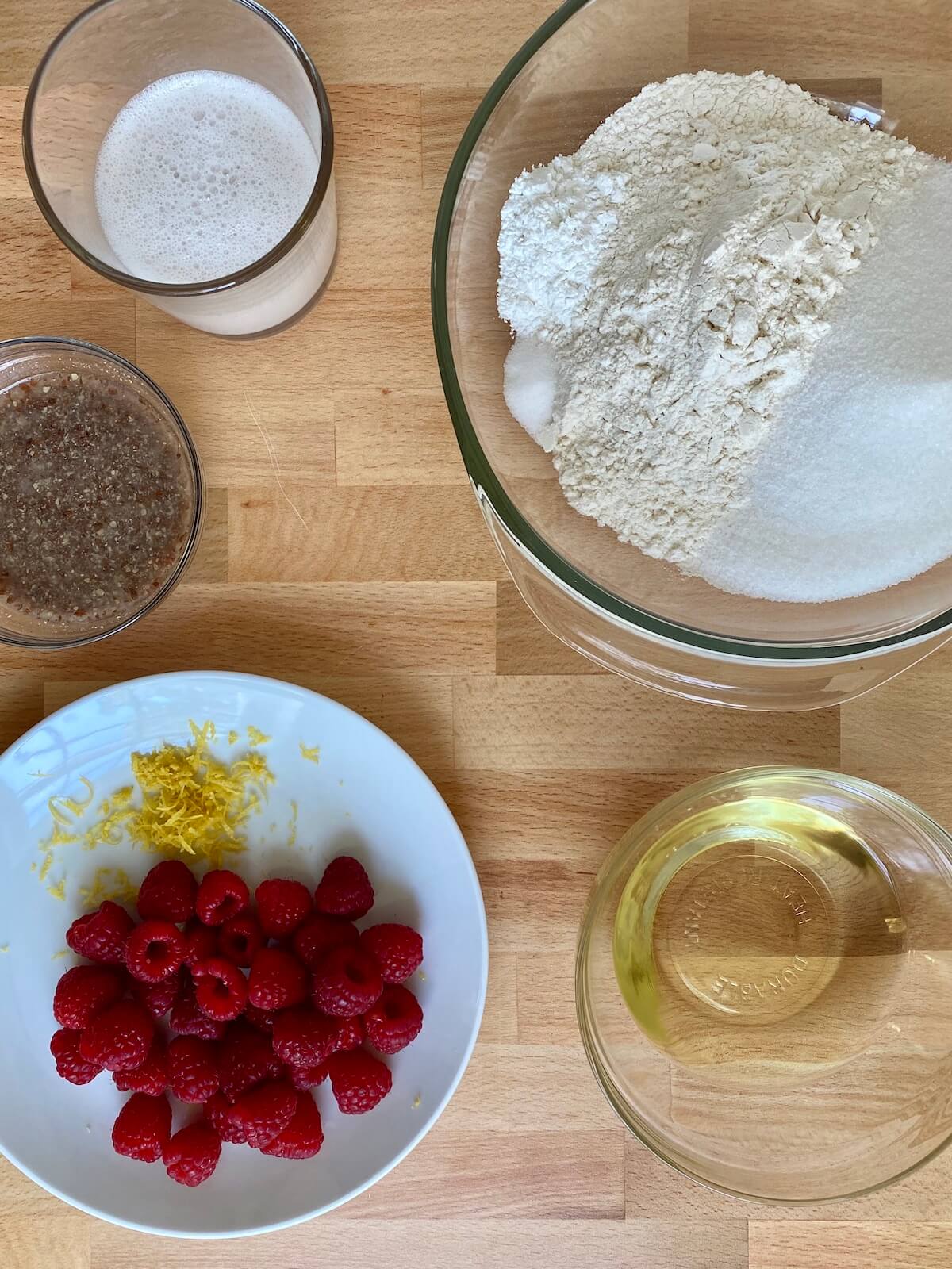 The ingredients to make the vegan raspberry muffins displayed in various bowls on a butcher block countertop.