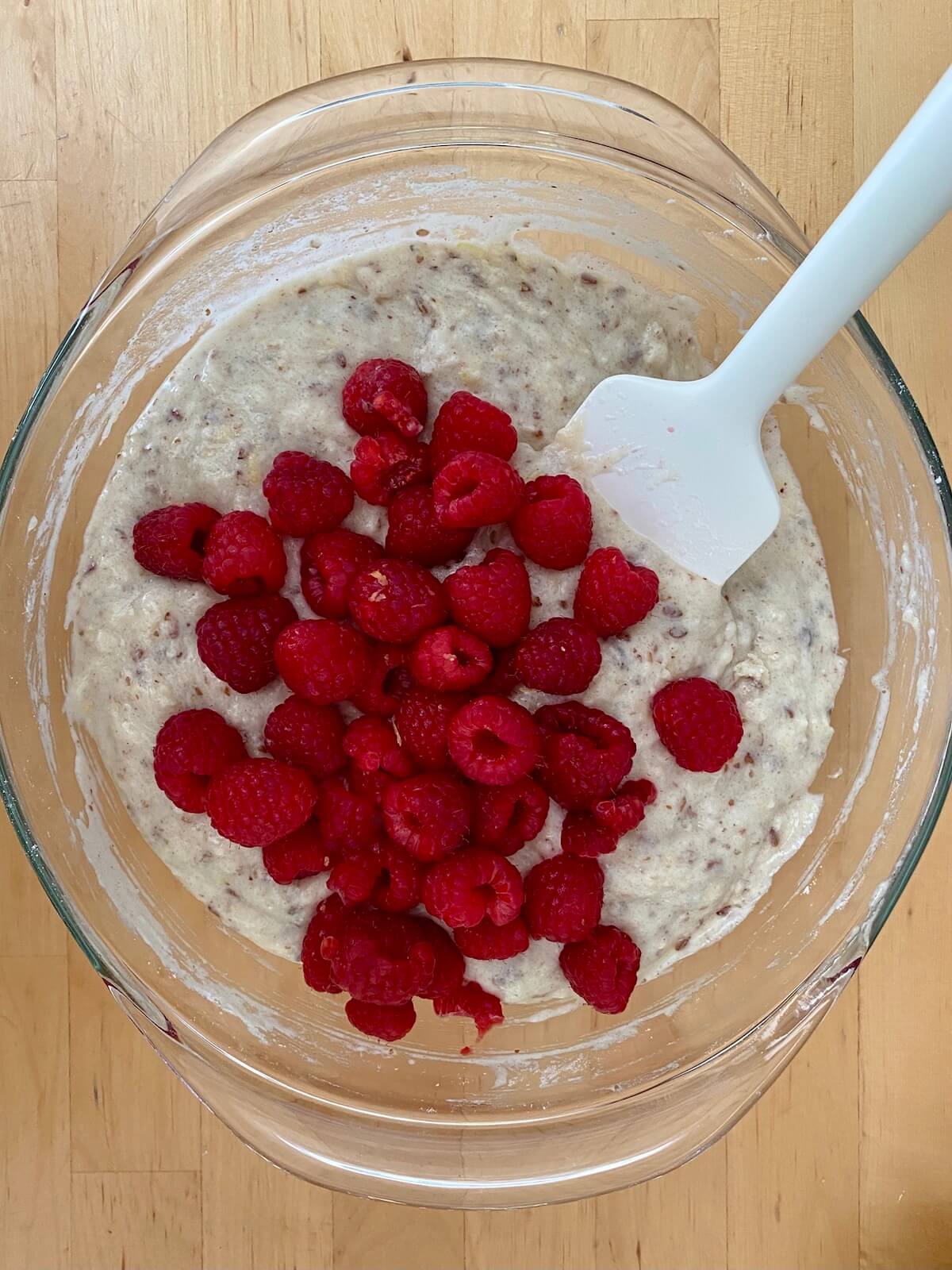 Fresh raspberries sitting on top of the mixed batter inside of a clear glass bowl. A rubber spatula is sticking out of the bowl to the right.