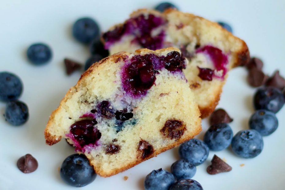 A blueberry chocolate chip muffin cut in half on a small white plate. There are fresh blueberries and chocolate chips scattered around the muffin on the plate.