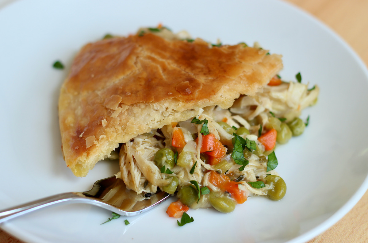 A slice of dutch oven chicken pot pie on a white plate. There is a fork under the chicken pot pie as if someone is about to take a bite. The dish is garnished with fresh chopped parsley.
