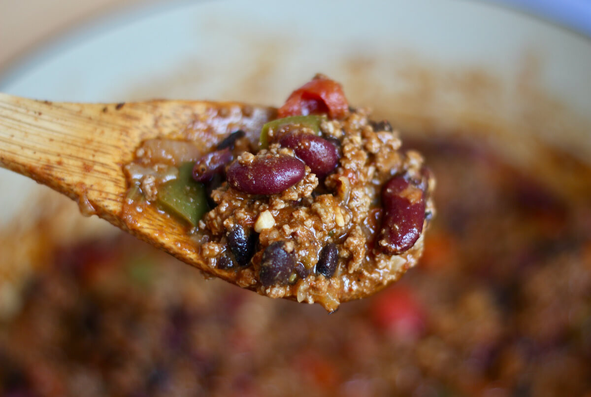 A wooden spoon holding up a scoop of chili over a large pot. The scoop of chili contains kidney beans, black beans, ground beef, tomatoes, and bell peppers.