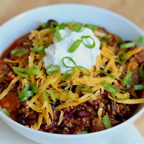 A white bowl filled with Dutch oven chili. The chili is topped with shredded cheddar cheese, sour cream, and scallions.