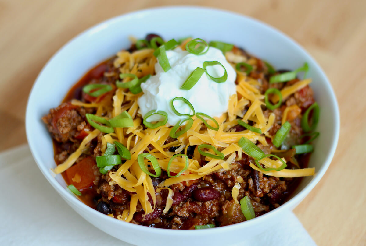 A white bowl of Dutch oven chili garnished with cheddar cheese, sour cream, and green onions.