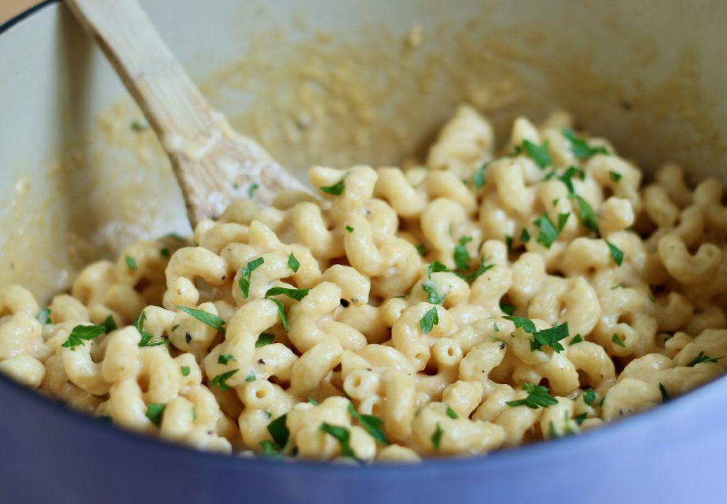 A lavender dutch oven is filled with one-pot mac and cheese. Out of focus in the background is a wooden spoon sticking out of the pot. The dish is garnished with fresh parsley.
