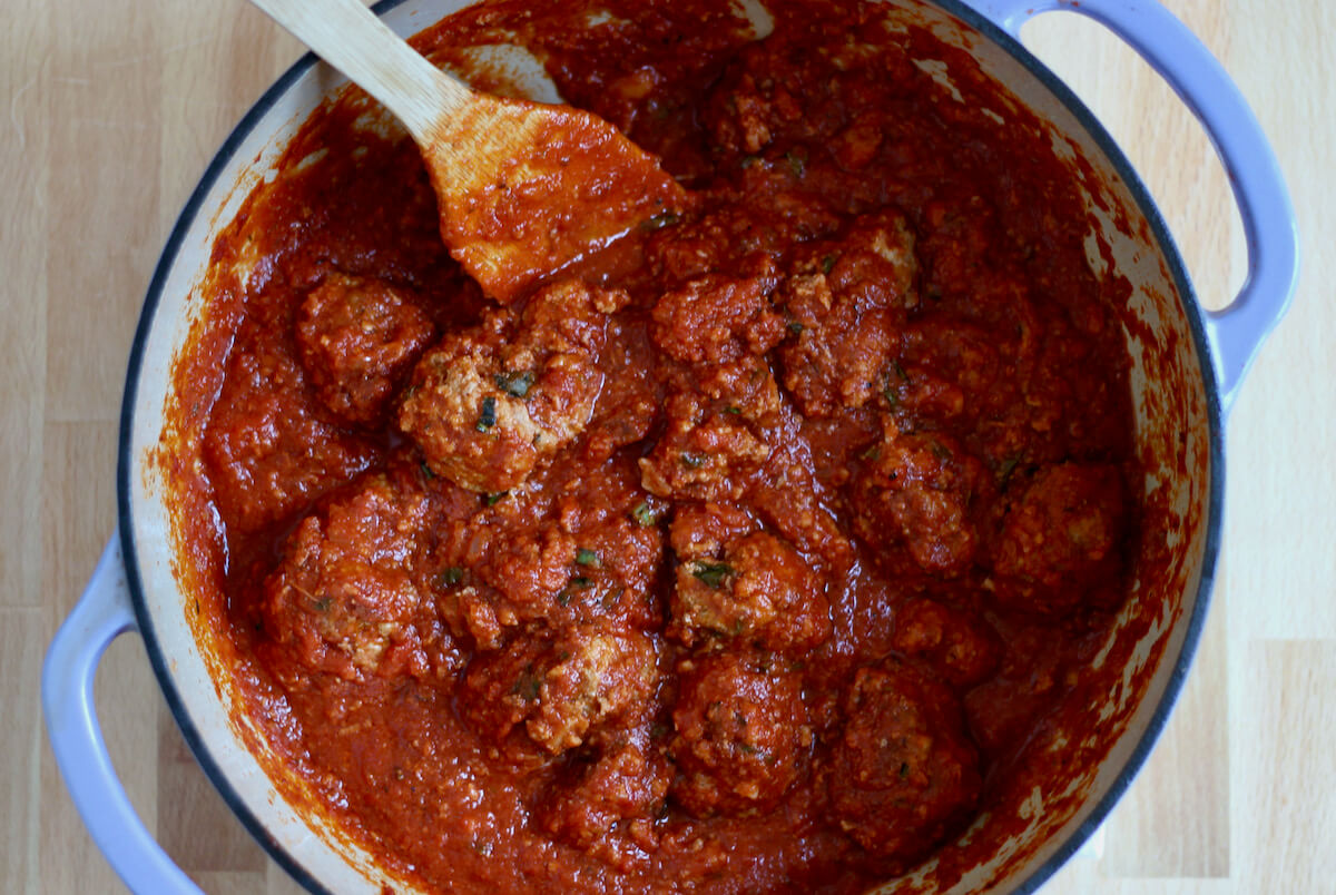 A large dutch oven filled with meatballs in tomato sauce. There is a wooden spoon sticking out of the pot to the left.