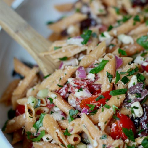 A large, white serving bowl filled with red lentil pasta salad. A wooden spoon is out of focus in the background, scooping some of the pasta salad.