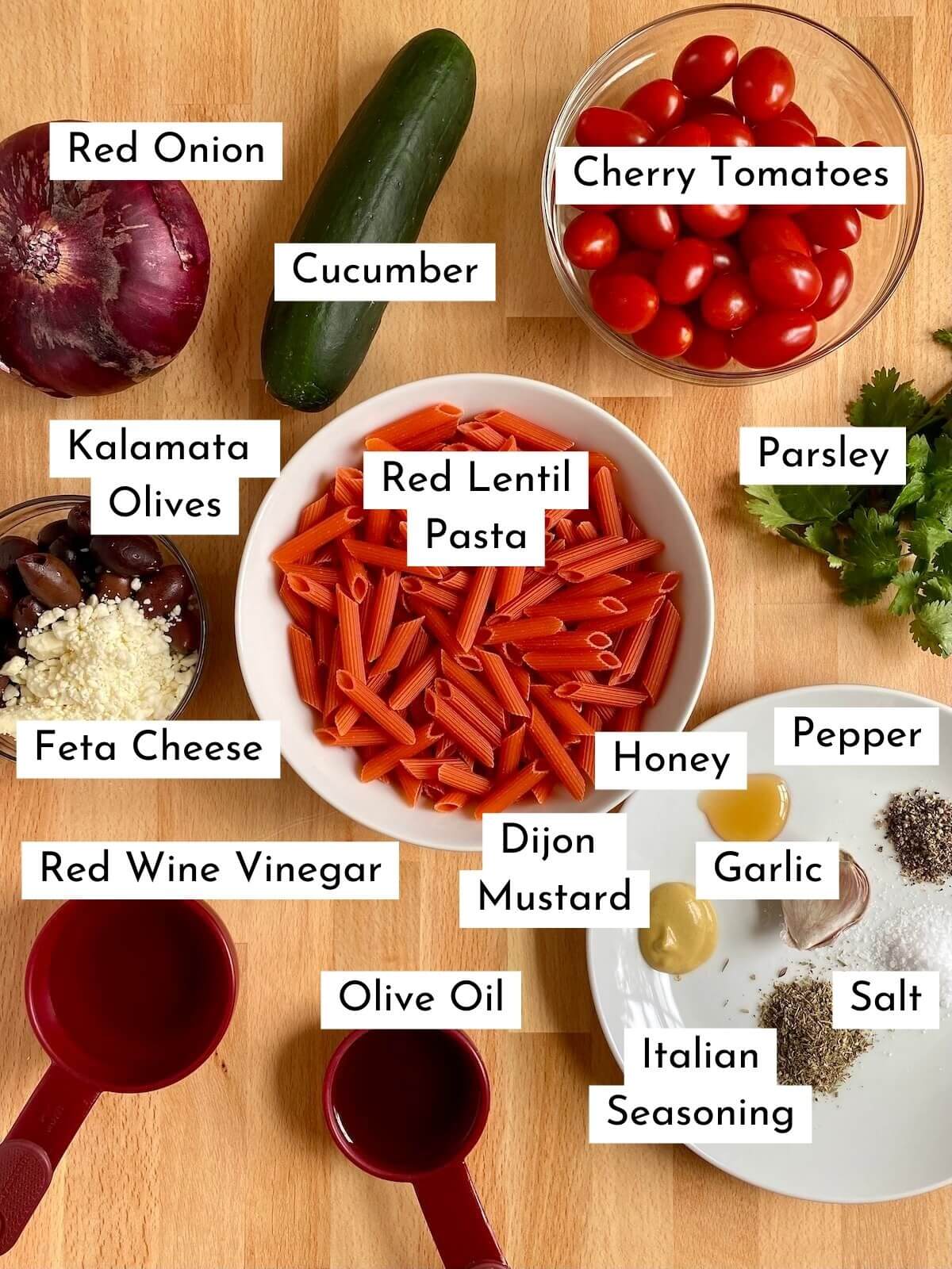 The ingredients to make red lentil pasta laid out on a butcher block countertop. There is text over each individual ingredient that states the name of each ingredient. The ingredients consist of red onion, cucumber, cherry tomatoes, Kalamata olives, feta cheese, red lentil pasta, parsley, red wine vinegar, olive oil, dijon mustard, honey, garlic, Italian seasoning, salt, and pepper.