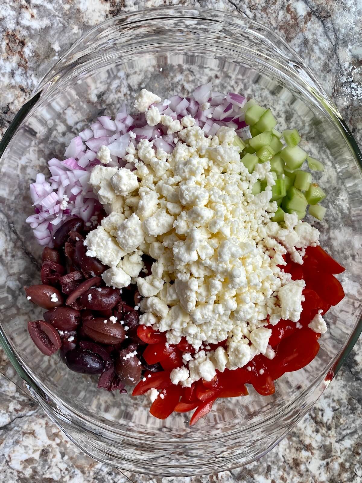 Chopped cucumber, red onion, cherry tomatoes, Kalamata olives, and feta cheese in a clear glass mixing bowl.