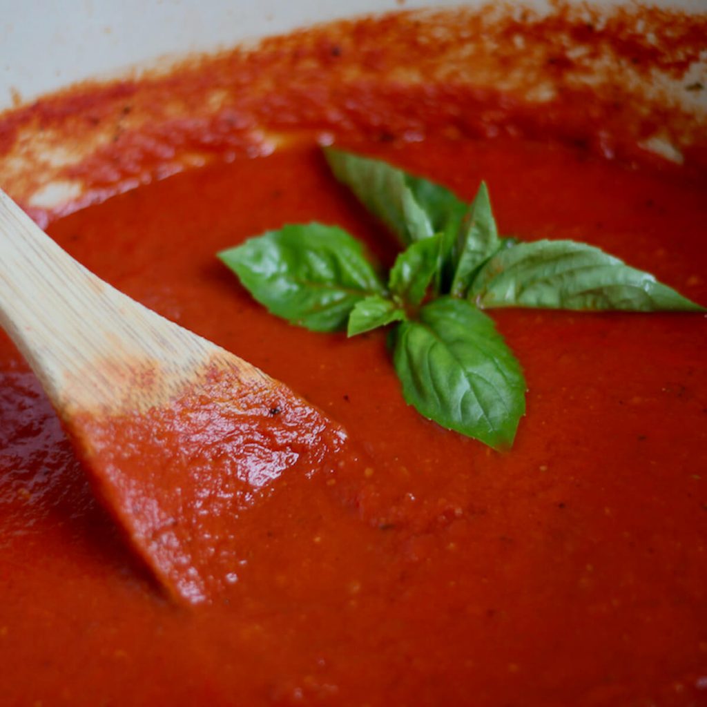 A Dutch oven filled with San Marzano tomato sauce. To the left, there is a wooden spoon sticking out of the sauce. There are a few basil leaves placed on top of the sauce as garnish.