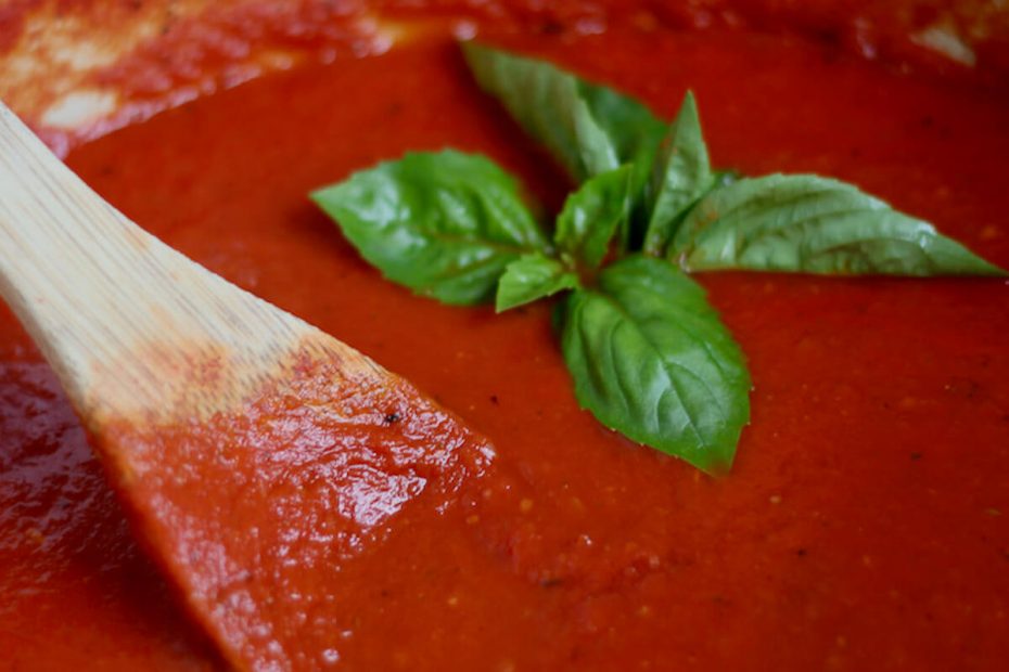 A Dutch oven filled with San Marzano tomato sauce. To the left, there is a wooden spoon sticking out of the sauce. There are a few basil leaves placed on top of the sauce as garnish.