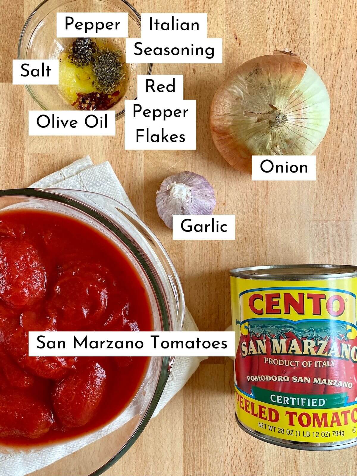 The ingredients to make San Marzano tomato sauce displayed on a butcher block countertop. There is text over each ingredient, stating what it is. The ingredients include San Marzano tomatoes, garlic, onion, olive oil, Italian seasoning, red pepper flakes, salt, and pepper.