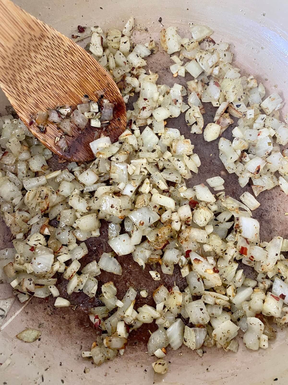 Onion, garlic, and seasonings being sautéed in a Dutch oven. There is a wooden spoon inside the pot, stirring the aromatics.
